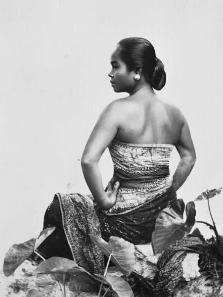 Historical portrait of a mother in traditional belly binding, seated among broad leaves.